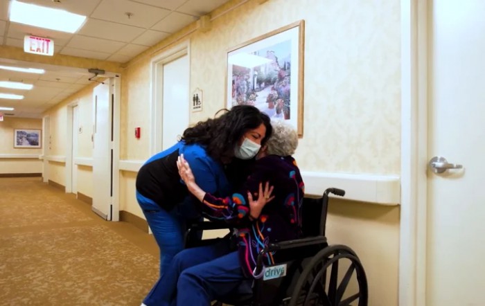 A caregiver wearing a mask hugs an elderly person in a wheelchair in a nursing home hallway.