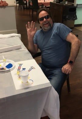 A person sitting at a table with art supplies, waving to the camera.