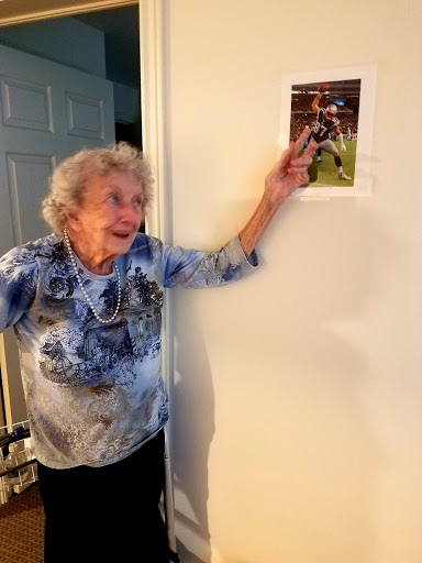 An elderly woman joyfully points at a photo displayed on the wall.