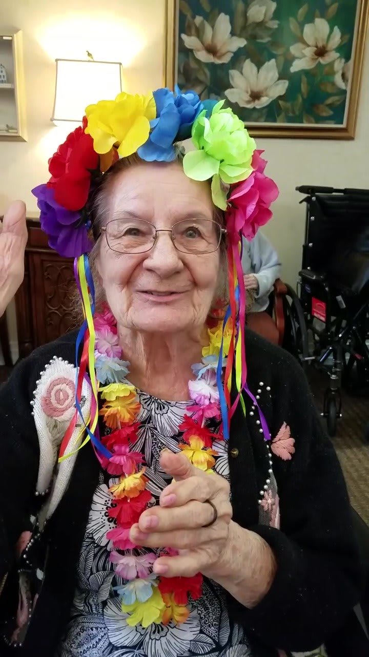 An elderly woman wearing a colorful flower crown and lei, smiling joyfully at an event.