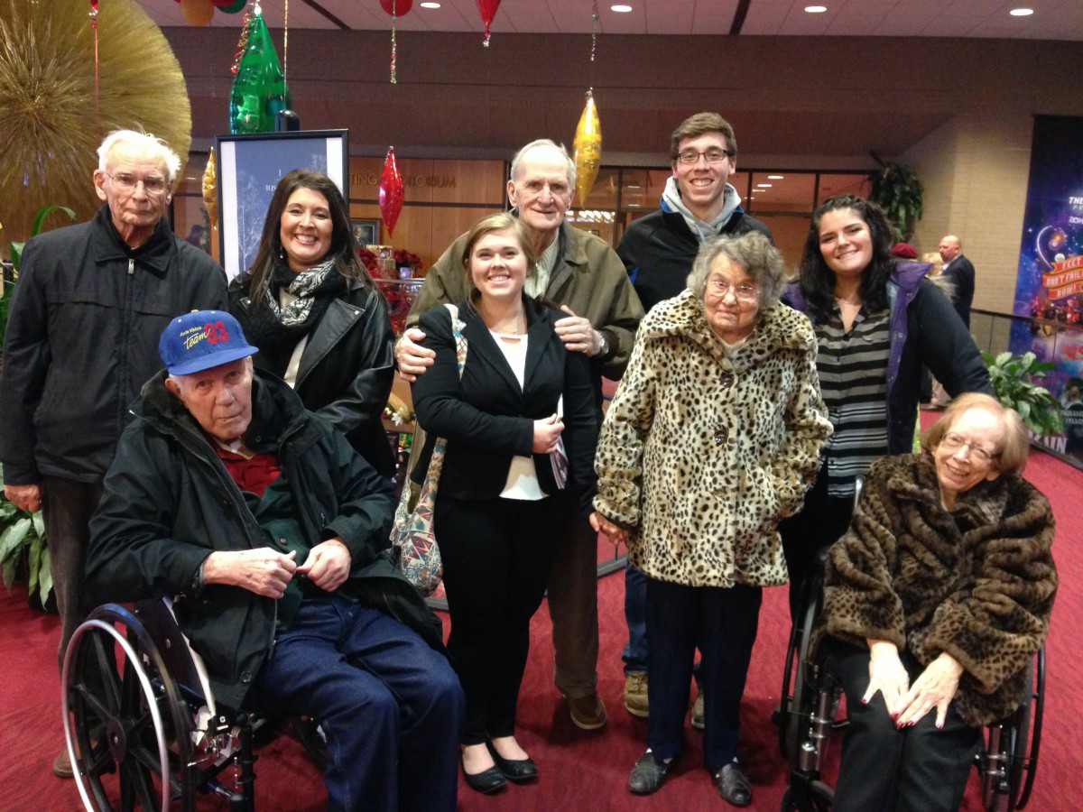 A group of people, including elderly individuals and some in wheelchairs, posing together indoors with smiling faces.