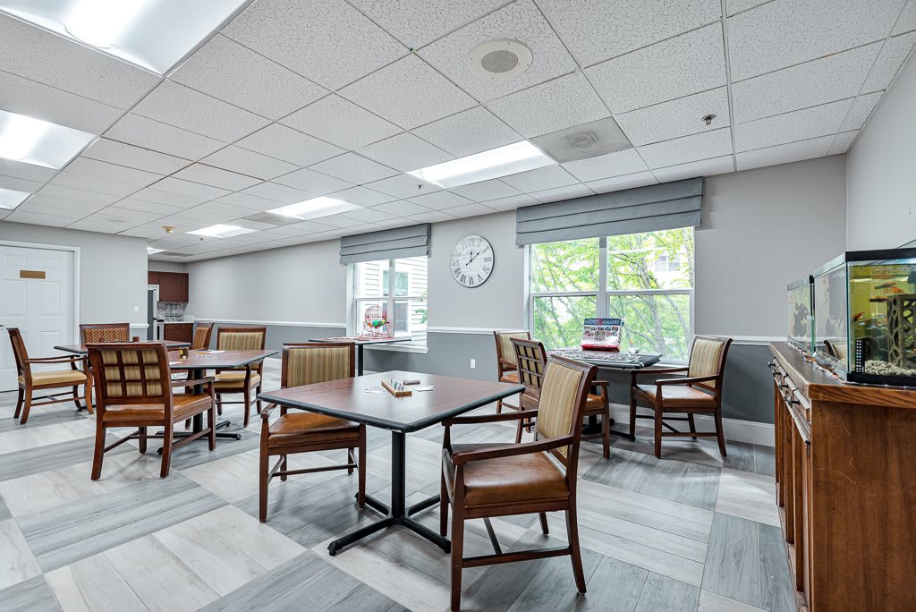 A bright and modern dining area with several tables and chairs, featuring large windows and a wall clock.