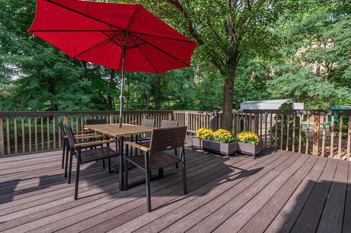 A serene outdoor patio with a wooden deck, featuring a dining table set under a red umbrella, surrounded by lush trees and potted flowers.