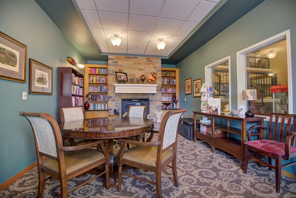 A cozy library with a fireplace, surrounded by bookshelves and a wooden table with chairs.