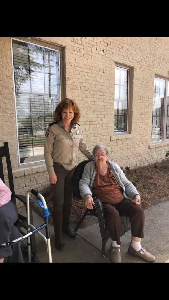 A caregiver standing next to an elderly person in a wheelchair outside a building.