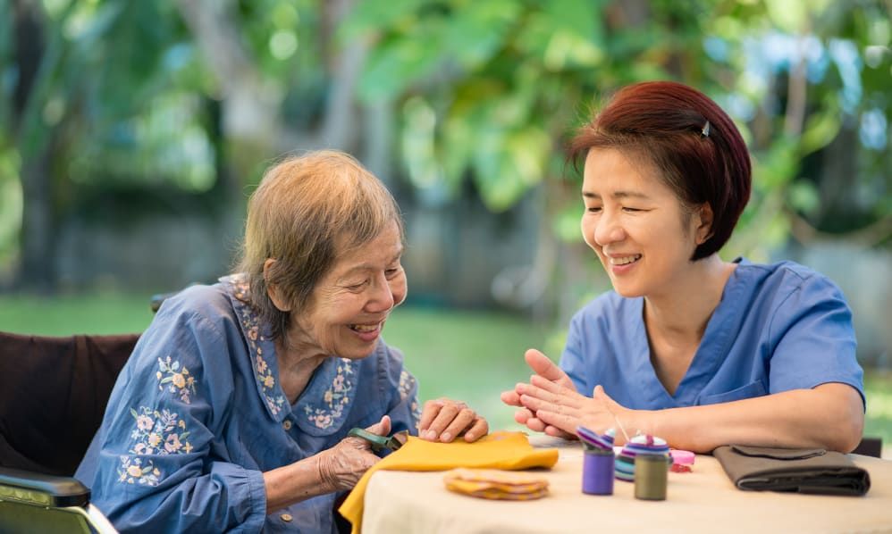 An elderly woman and a younger woman enjoy sewing together outdoors, sharing smiles and companionship.