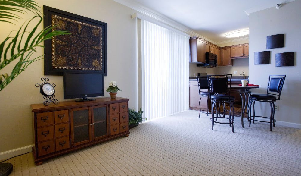 A cozy living room with a TV stand, leading into an open kitchen with bar stools and natural light from large window blinds.