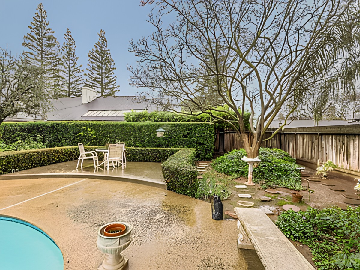 A serene backyard patio area with outdoor seating, surrounded by lush hedges and a tree.