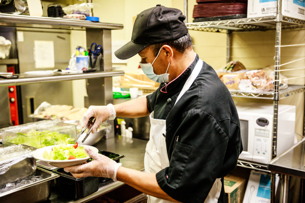A chef wearing a mask and gloves prepares a dish in a restaurant kitchen.