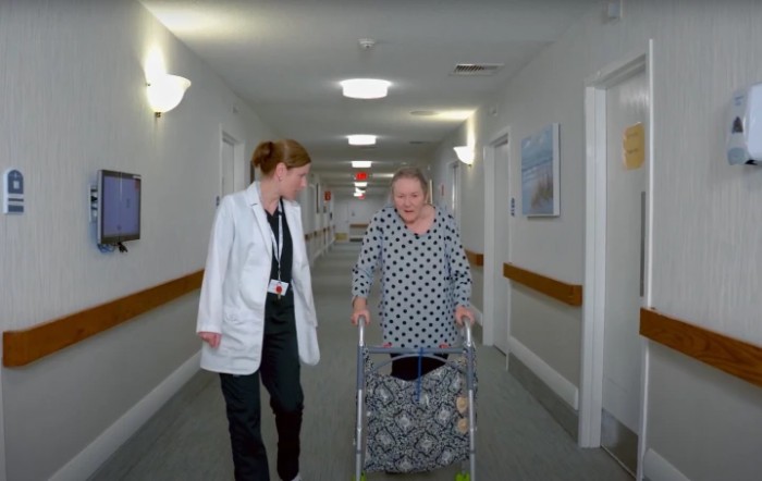 A doctor walks alongside an elderly patient using a walker in a hospital hallway.