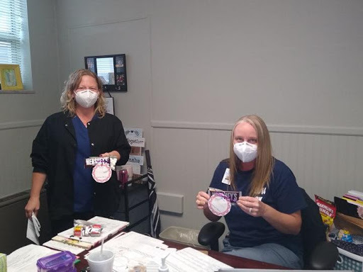 Two women in an office setting, wearing masks and holding up items while sitting at a desk.