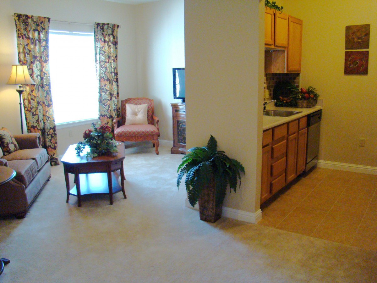A cozy living room with a view into a small kitchen, featuring warm lighting and comfortable furniture.