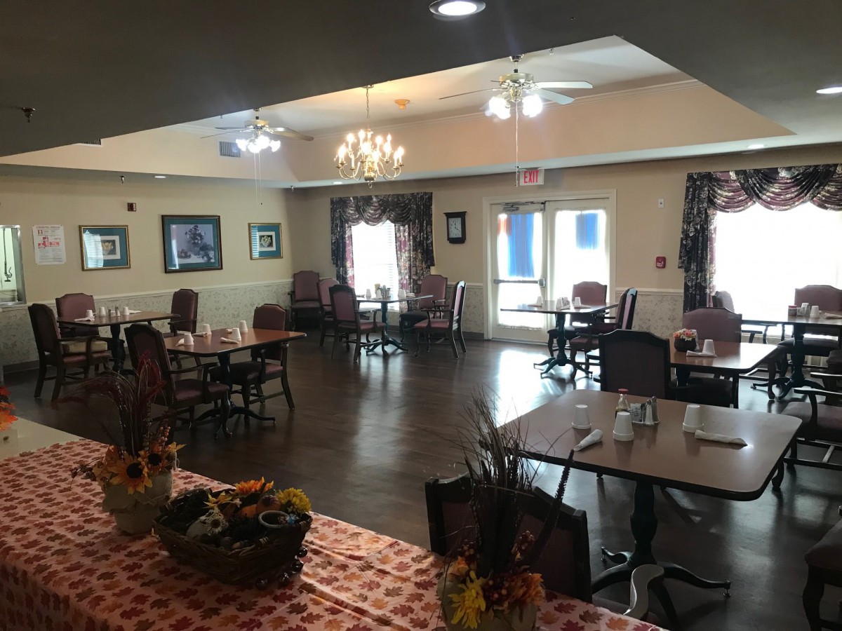 A cozy dining room with neatly arranged tables and chairs under a chandelier, featuring large windows and decorative elements.