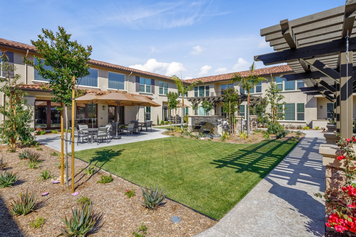 A sunny courtyard featuring a landscaped garden and outdoor seating area, surrounded by a residential building.