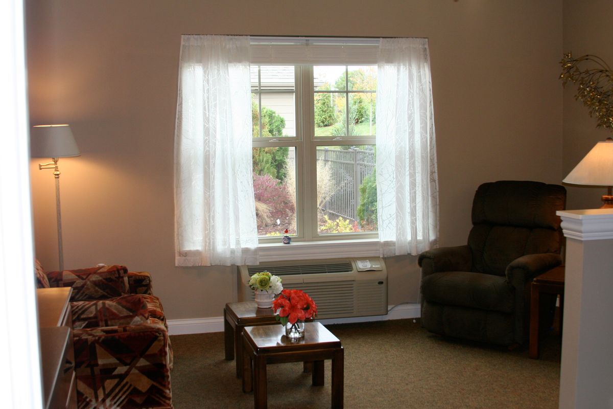 A cozy living room with a window adorned with sheer curtains, featuring an armchair, sofa, and a small table.