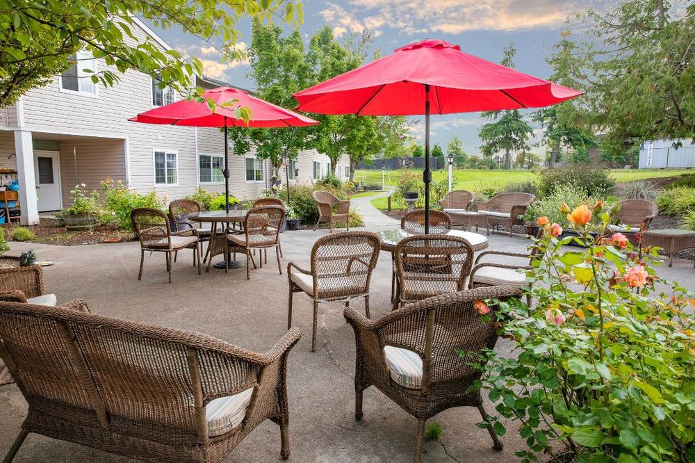 A cozy patio with rattan furniture and red umbrellas, surrounded by a lush garden.