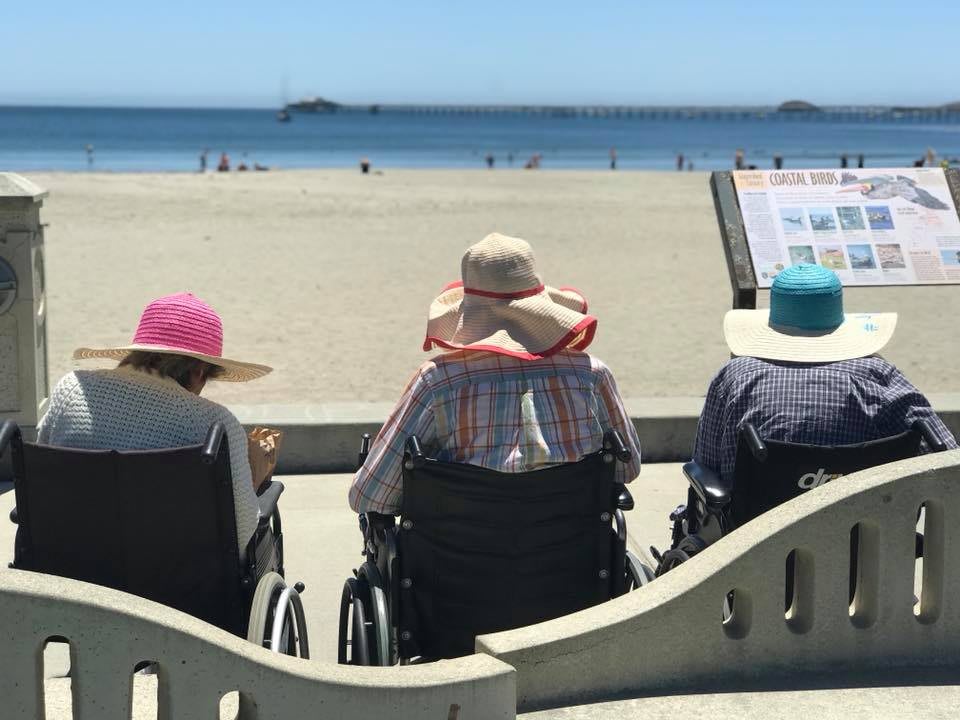 Three people wearing hats sit in wheelchairs, enjoying a view of the beach and ocean.
