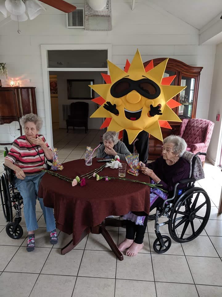 Elderly individuals sitting around a table, joined by someone in a sun costume.