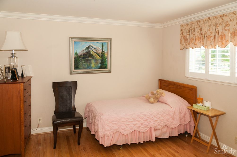 A cozy bedroom with a single bed dressed in pink, a teddy bear on the bed, and a painting hanging above. A dark chair sits next to the bed near a window with curtains.
