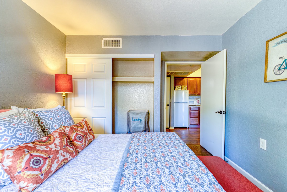 A cozy bedroom featuring a bed with patterned bedding, a lamp on the side, and an open closet. The room opens into a hallway leading to the kitchen.