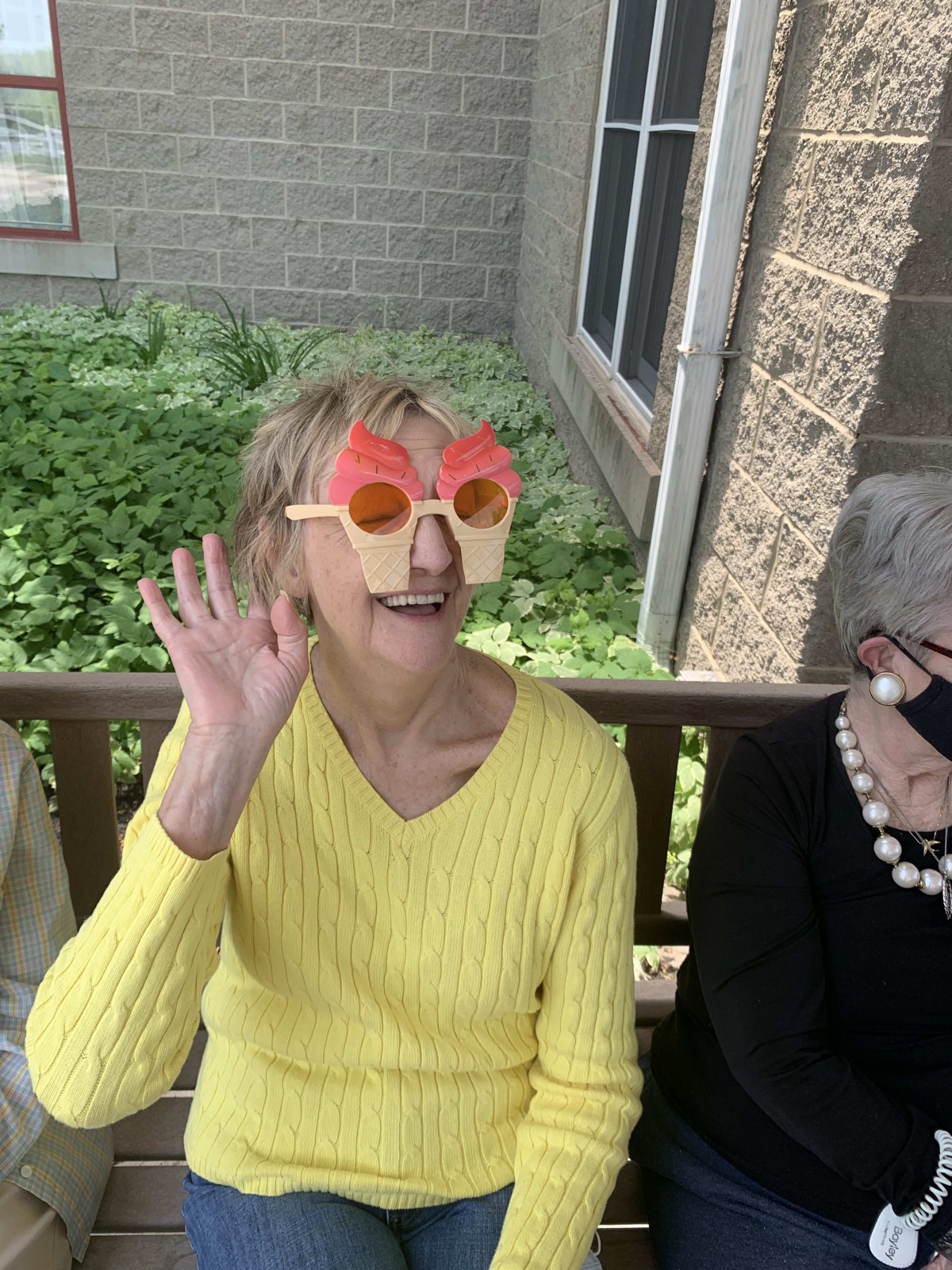 A woman in a yellow sweater wearing playful sunglasses waves while sitting on a bench outdoors.