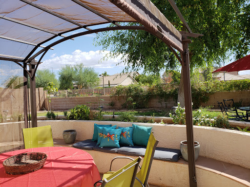 A cozy outdoor patio featuring a colorful table and comfortable seating under a sunshade, surrounded by greenery.
