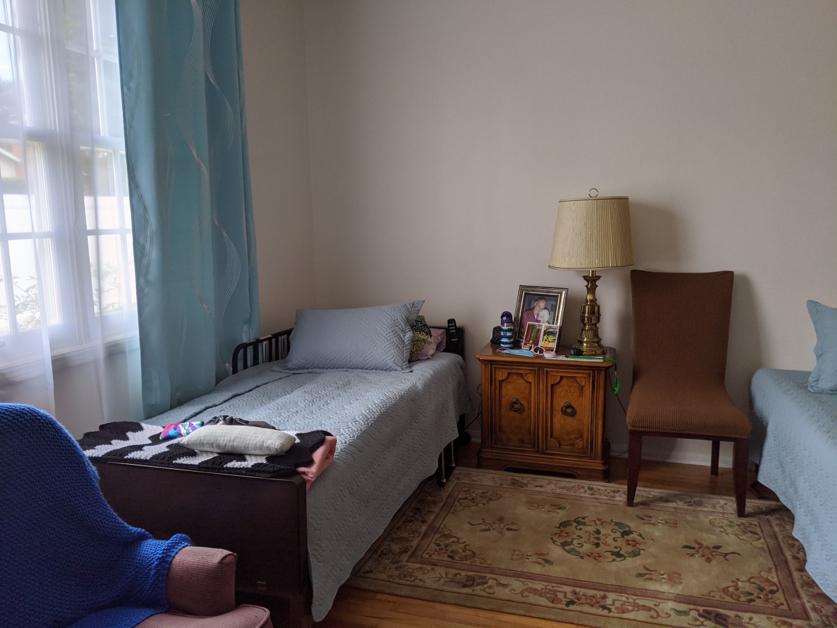 A cozy bedroom featuring a single bed, a vintage lamp on a wooden nightstand, and a chair near the window with blue curtains.