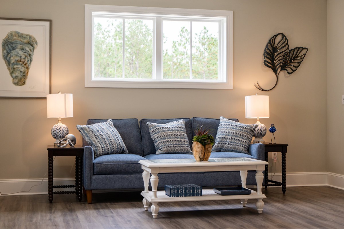 A cozy living room with a blue sofa, stylish cushions, and two lamps on either side of the couch. A white coffee table stands in front of the sofa, all set against a light-colored wall with a window above.