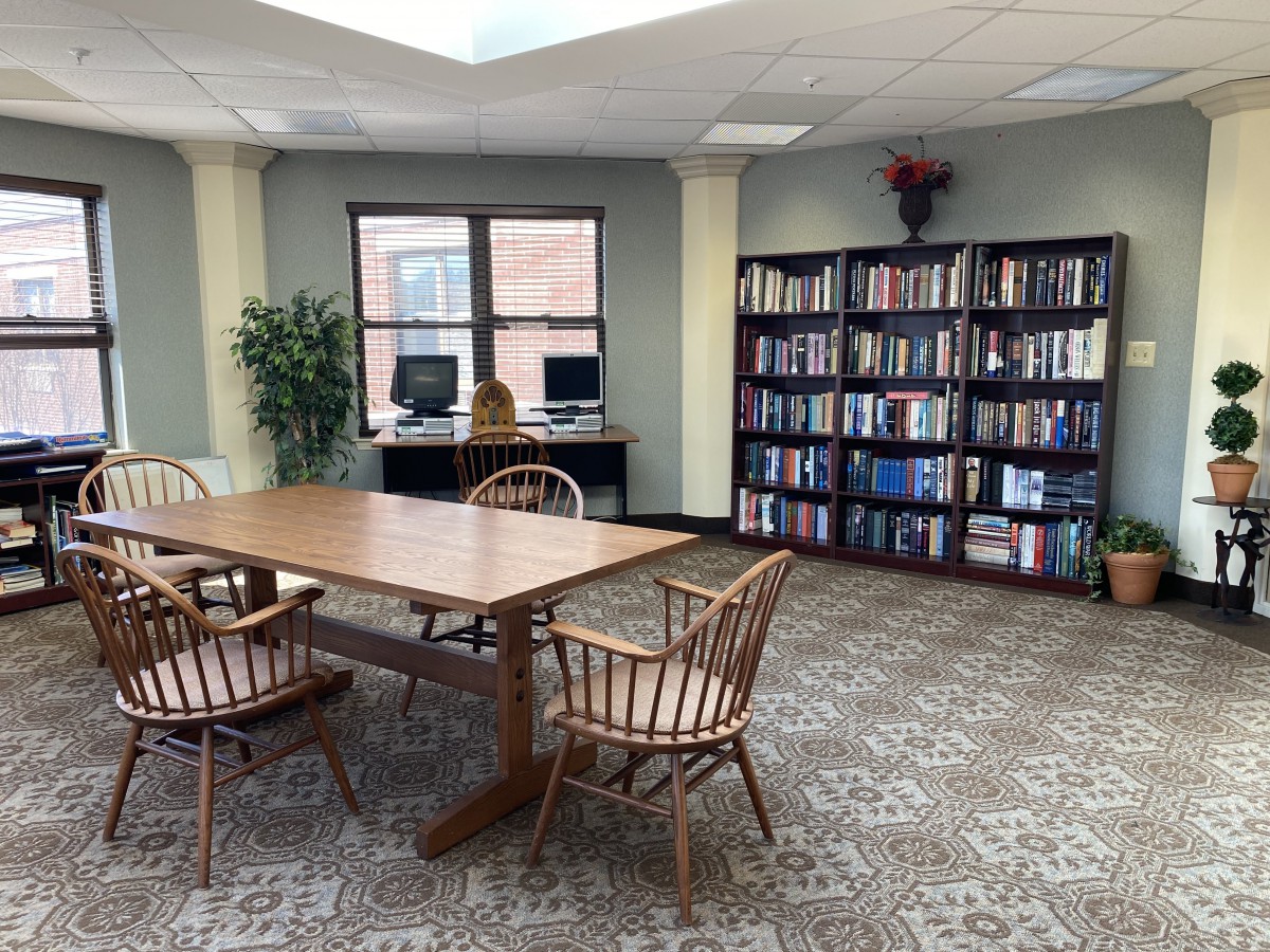 A quiet library space with a wooden table and chairs, bookshelves lined with books, and computers by the window.