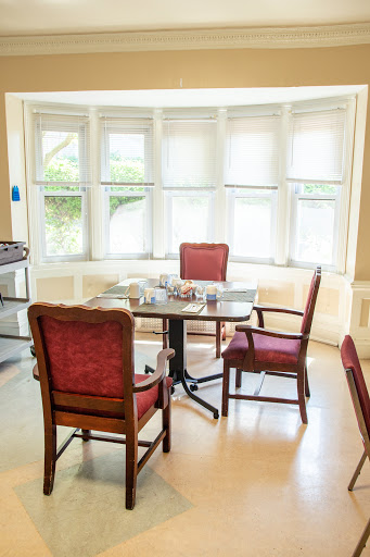 A cozy dining area featuring a round table surrounded by red upholstered chairs, illuminated by natural light from large windows.