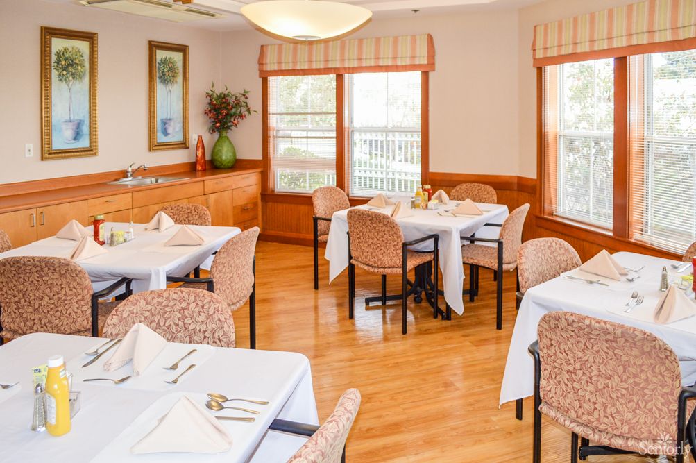 A cozy dining room with neatly arranged tables and chairs, featuring a wooden floor and natural lighting from large windows.
