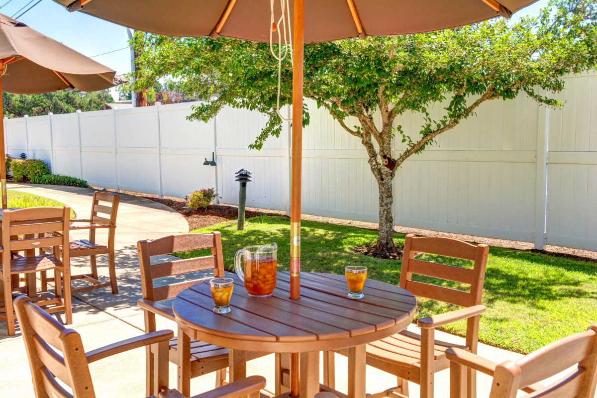 A sunny patio with wooden outdoor furniture, including a table and chairs under an umbrella, featuring a pitcher of lemonade.