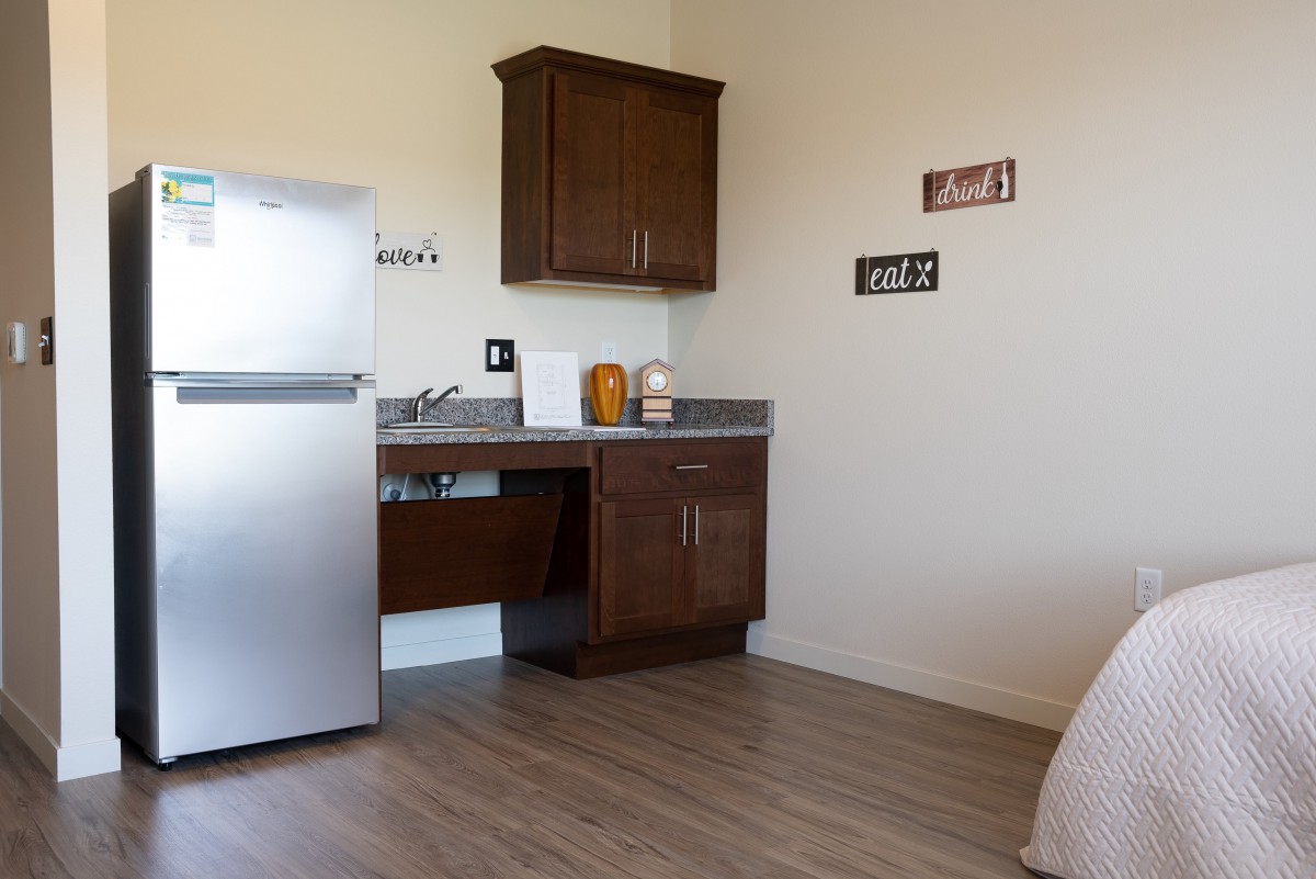 A simple kitchen area with a fridge, cabinets, and a sink on wooden flooring.