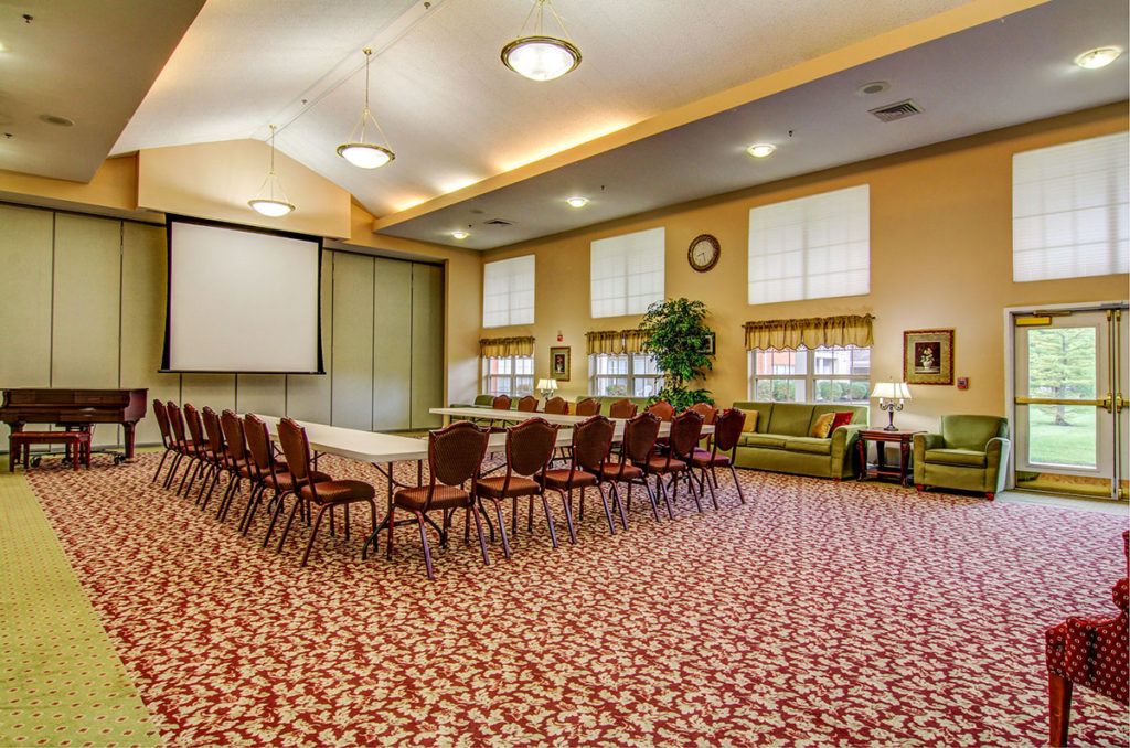 A well-lit conference room with arranged chairs and tables, featuring a projector screen and carpeted flooring.
