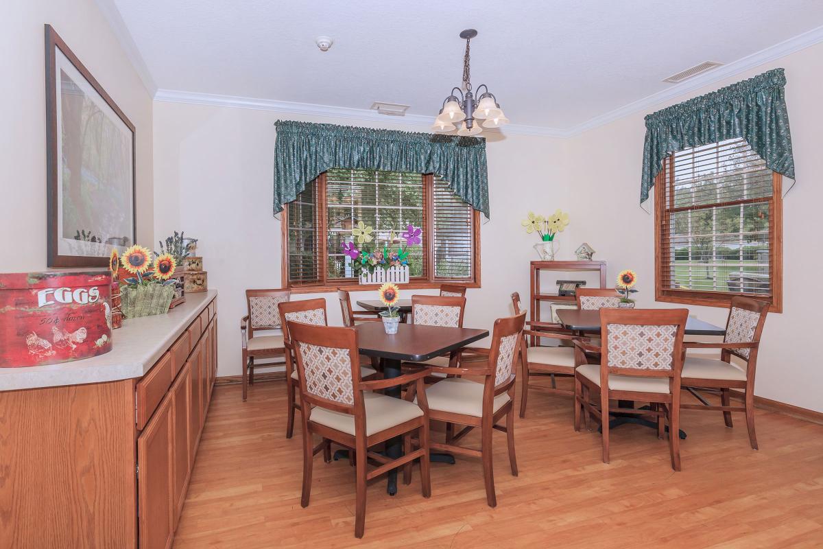 A cozy dining room with wooden flooring, featuring a central table surrounded by chairs and adorned with floral decorations and window curtains.