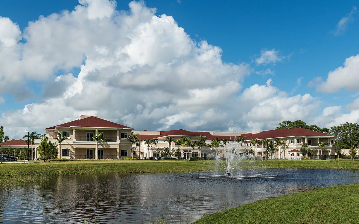 A picturesque view of buildings with red-tiled roofs reflected in a serene lake, featuring a central fountain under a partly cloudy sky.