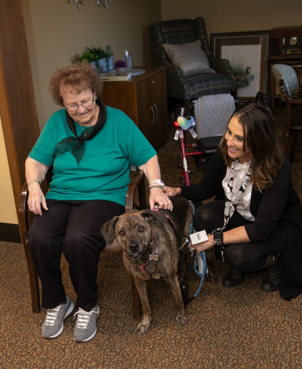 An elderly woman and a younger woman enjoying the company of a dog indoors.