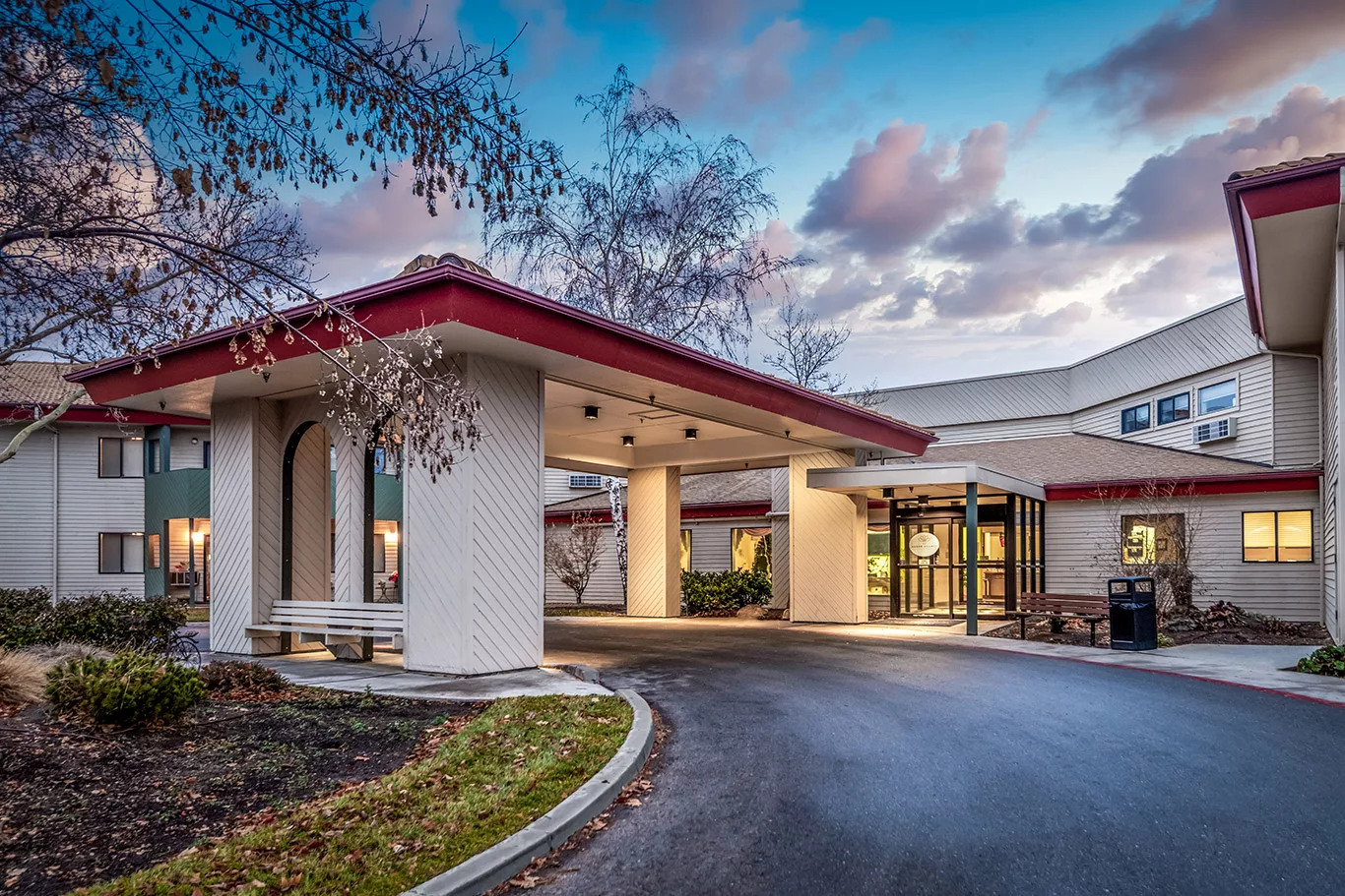 A modern hotel entrance with a driveway and landscaped surroundings during sunset.