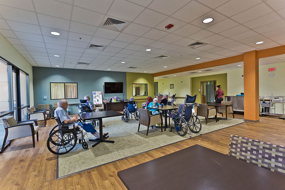 A common area in a nursing home with residents sitting in wheelchairs and chairs, surrounded by warm-toned decor.