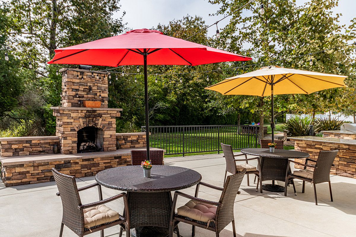 A cozy outdoor patio area featuring tables with chairs and colorful umbrellas, set against a backdrop of trees and a brick fireplace.