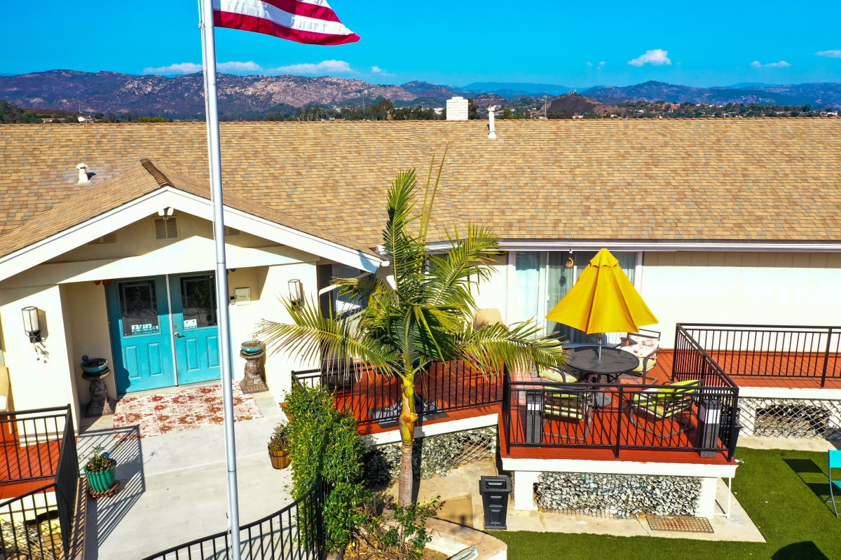 A backyard view featuring a patio with outdoor furniture, an American flag, and mountains in the background on a sunny day.