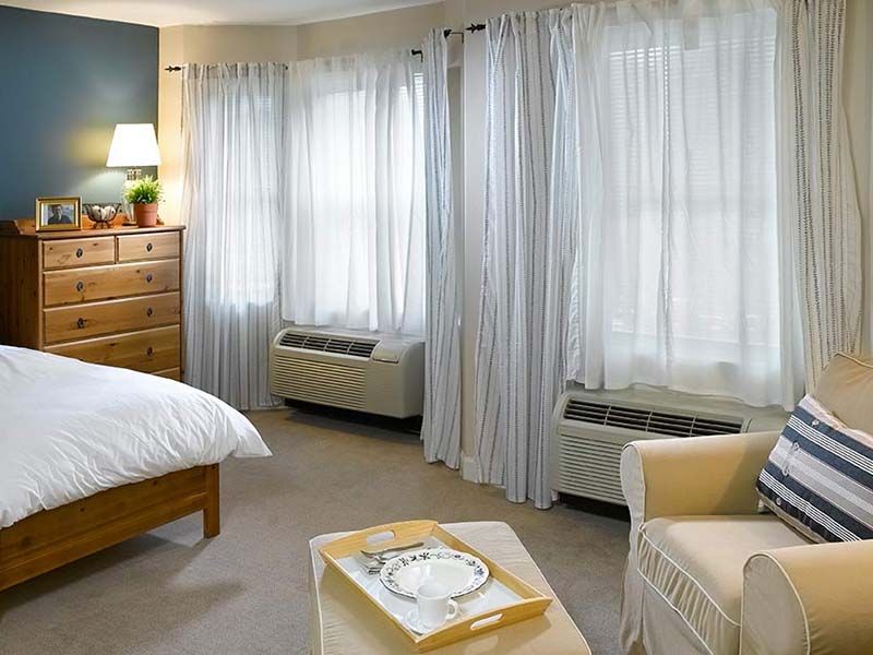 A cozy bedroom with large windows, sheer curtains, a comfortable chair, and a wooden dresser.