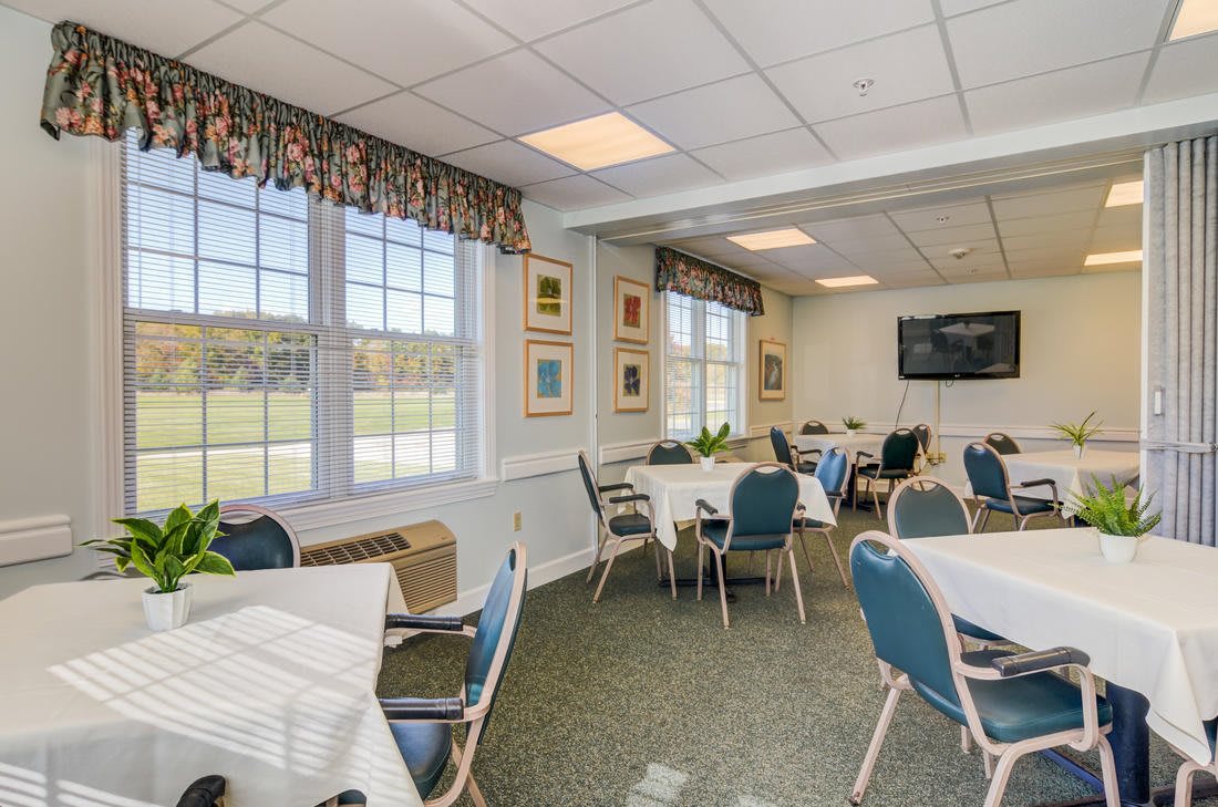 A well-lit dining room with tables and chairs, large windows, and a television on the wall.