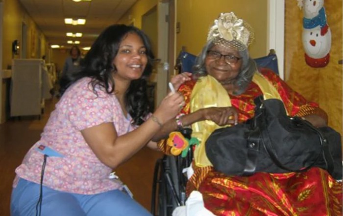 A healthcare worker smiling next to an elderly woman in a wheelchair, dressed for a celebration.