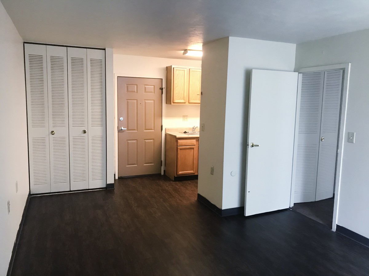 An empty apartment interior with dark hardwood floors and a small kitchen area.