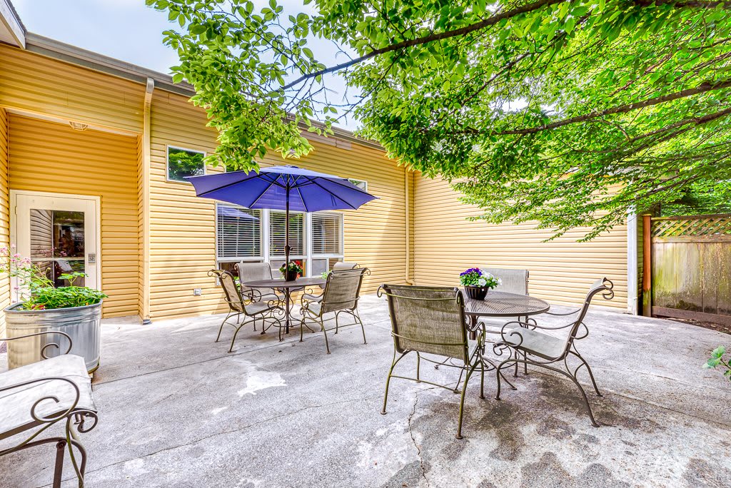A cozy backyard patio with tables, chairs, and a blue umbrella under the shade of a tree.