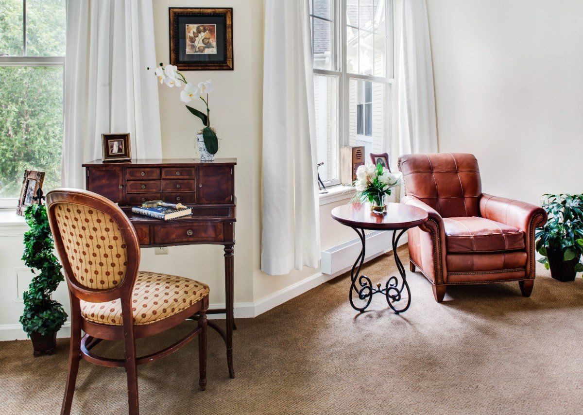 A cozy living room with elegant furniture, featuring a chair, small desk, and side table near a window.