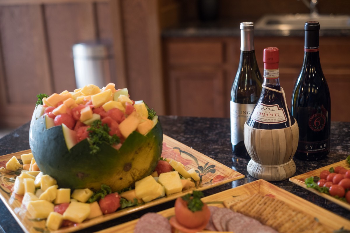 An elegant fruit arrangement beside two bottles of wine on a buffet table.