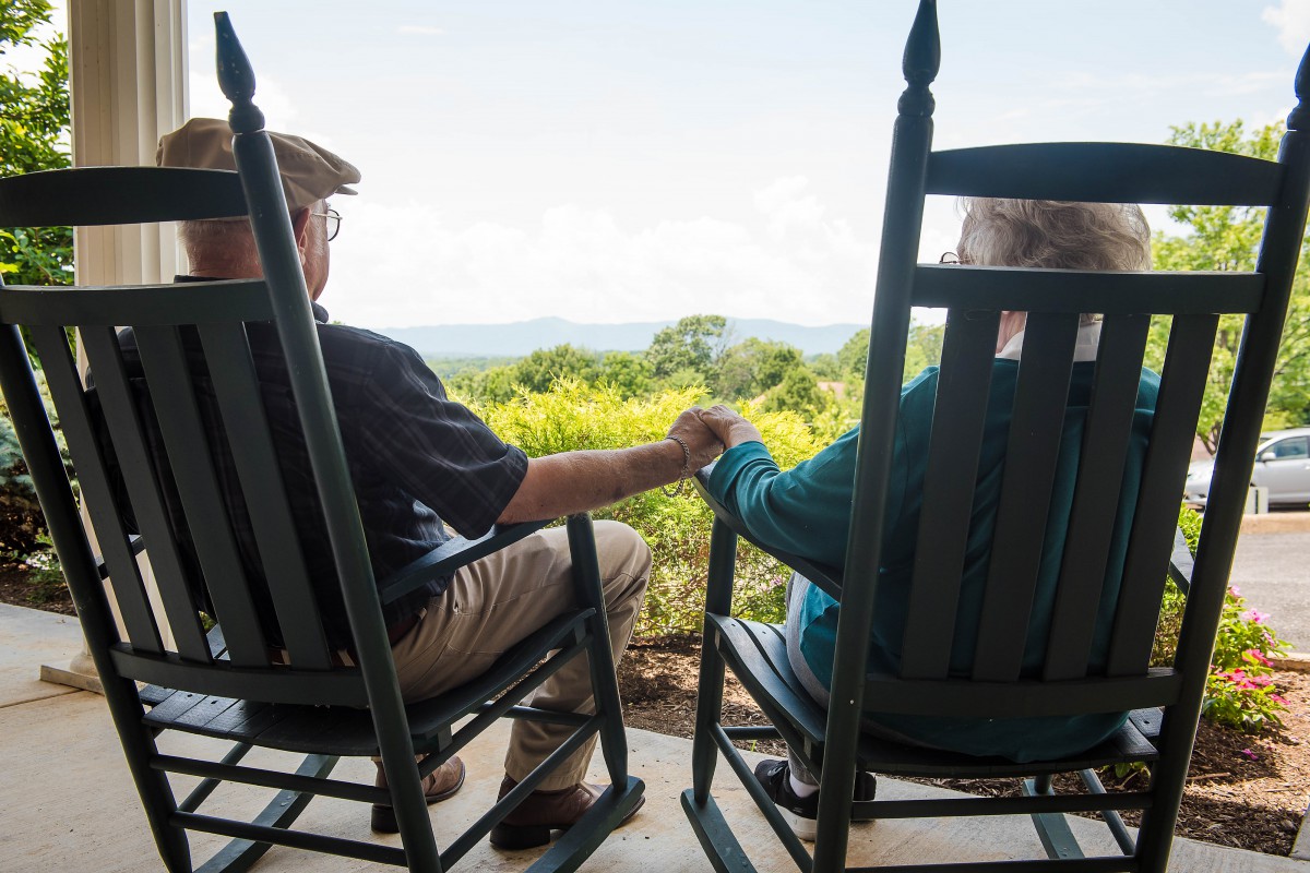 An elderly couple sits on rocking chairs, holding hands and enjoying a scenic view from their porch.
