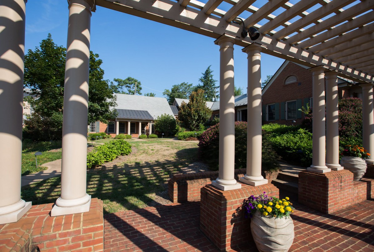 A serene outdoor area with a pergola and columns, surrounded by lush gardens and brick pathways.